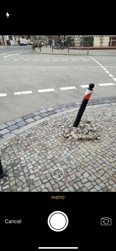 The mobile app's in-app camera capturing a photo of a damaged bollard leaning out of cobblestone pavers on a street corner.